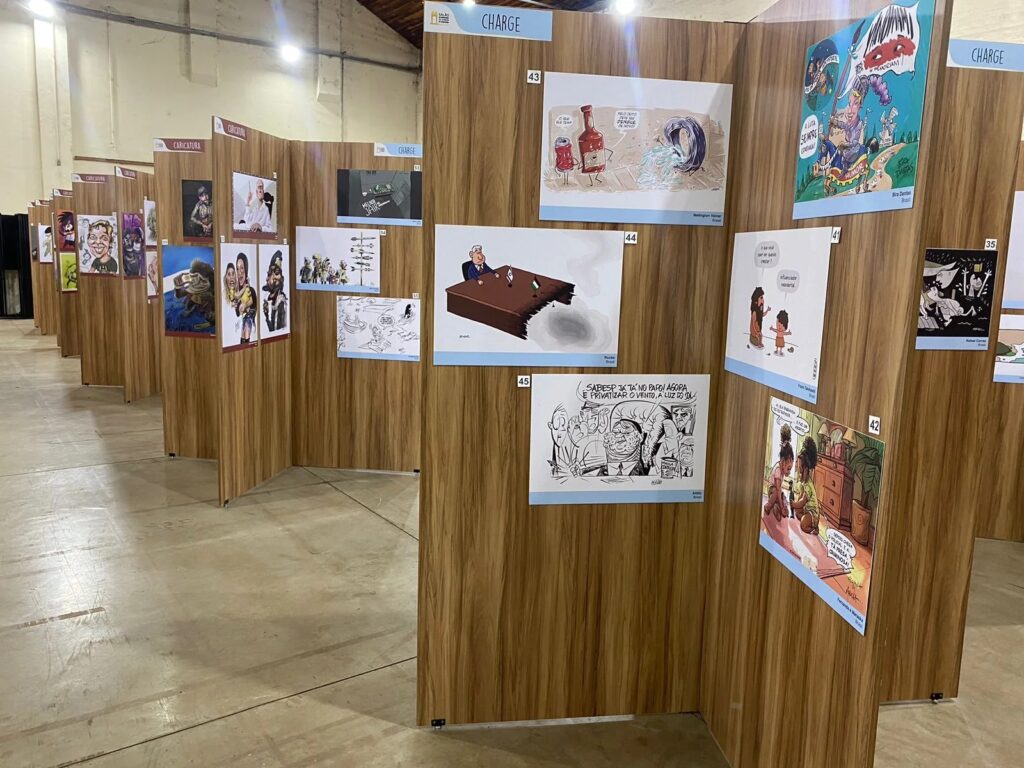 A imagem exibe uma exposição de caricaturas e charges. As obras estão montadas em painéis de madeira, criando um ambiente de galeria. Várias peças são visíveis, cada uma com um número e algumas com o nome do artista e seu país. Do lado direito da imagem, um painel apresenta várias obras numeradas: - A obra rotulada como "43" mostra uma garrafa de ketchup e uma lata de refrigerante, ambas com características antropomórficas, em uma cena que parece envolver encanamento. O texto "Wellington Viana Brasil" está visível abaixo. - Abaixo da "43", a obra "44" retrata uma pessoa sentada à mesa, que está parcialmente quebrada. O nome "Rucke Brasil" é visível. - Mais abaixo, a obra "45" é um desenho em preto e branco de várias pessoas, com texto acima delas. O nome "Affonso Brasil" aparece.