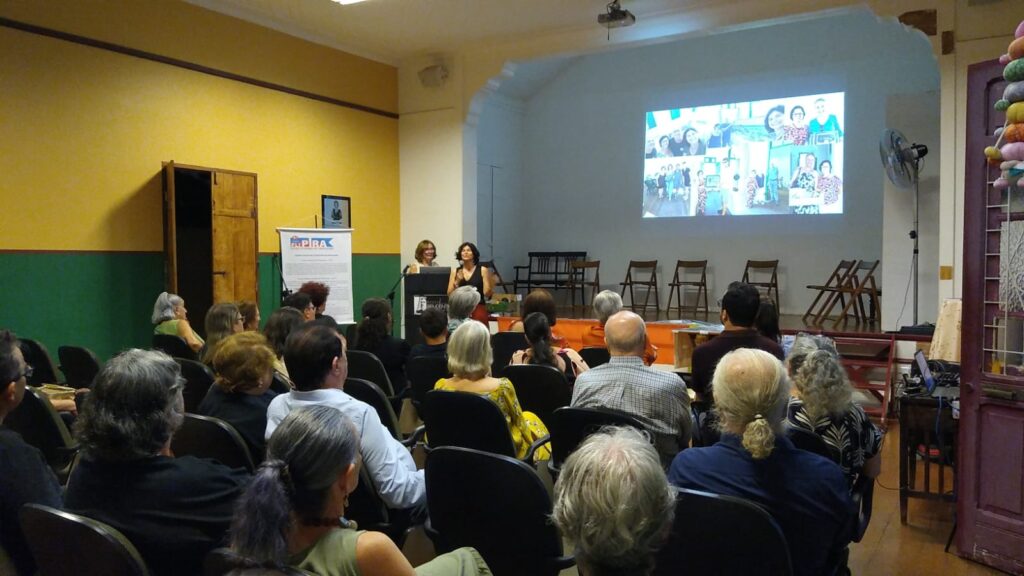 A imagem retrata uma sala de eventos com um público sentado em cadeiras, voltado para um palco onde duas mulheres estão apresentando. Uma delas está em pé, gesticulando enquanto fala, e a outra está sentada atrás de um púlpito.Ao fundo, um telão exibe uma colagem de fotos, possivelmente de eventos passados ou pessoas relacionadas ao tema da apresentação. A sala tem paredes em tons de amarelo e verde, e há uma porta de madeira à esquerda. Cadeiras de madeira dobráveis estão dispostas atrás das apresentadoras.A iluminação sugere um ambiente interno, possivelmente uma auditório ou sala de conferências. O público, composto por pessoas de diversas idades, parece atento à apresentação.