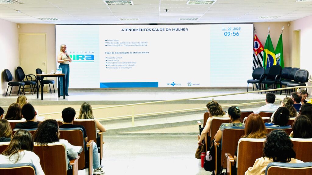A imagem mostra uma palestra em uma sala, com uma apresentadora em pé ao lado de uma grande tela. A tela exibe o título "ATENDIMENTOS SAÚDE DA MULHER" e uma lista de tópicos relacionados, como "Enfermeiros", "Médicos da estratégia saúde da família", "Ginecologistas/Equipe multiprofissional" e "Papel do Ginecologista na atenção básica". Abaixo, há logotipos do SUS, Saúde e Campinas. A data e hora "11-09-2025 09:56" também são visíveis na tela. A apresentadora, uma mulher loira vestindo uma blusa bege e calças azuis, está com um microfone na mão e parece estar falando com a audiência. Na frente da tela, há uma mesa com garrafas de água e papéis. A audiência, composta por homens e mulheres, está sentada em fileiras de cadeiras de madeira, olhando para a tela. Há bandeiras do estado de São Paulo, do Brasil e possivelmente de outro município atrás da apresentadora. A sala parece ser um auditório ou sala de conferências, com iluminação artificial vinda do teto.