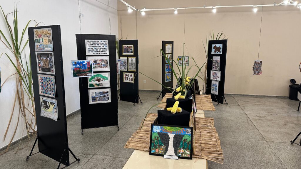 A imagem exibe uma exposição de arte em um ambiente interno, possivelmente uma galeria ou escola.No primeiro plano, à esquerda, há um painel preto vertical com várias obras de arte penduradas, incluindo desenhos, colagens e fotografias. Ao lado deste painel, altas hastes de plantas, possivelmente cana-de-açúcar, adicionam um elemento natural à cena.Em direção ao centro e à direita, outros painéis pretos semelhantes exibem mais trabalhos artísticos. Entre os painéis, em uma mesa coberta com esteiras de palha, há espigas de milho dispostas sobre bases pretas, criando uma instalação que combina arte e elementos agrícolas. Uma pintura vibrante de uma cachoeira em uma floresta está em destaque sobre as esteiras.O fundo é uma parede clara, iluminada por spots de luz no teto, que destacam as exposições. A disposição dos painéis e da instalação central sugere um evento cultural ou educacional, com foco em artes visuais e possivelmente em temas relacionados à natureza e à agricultura.