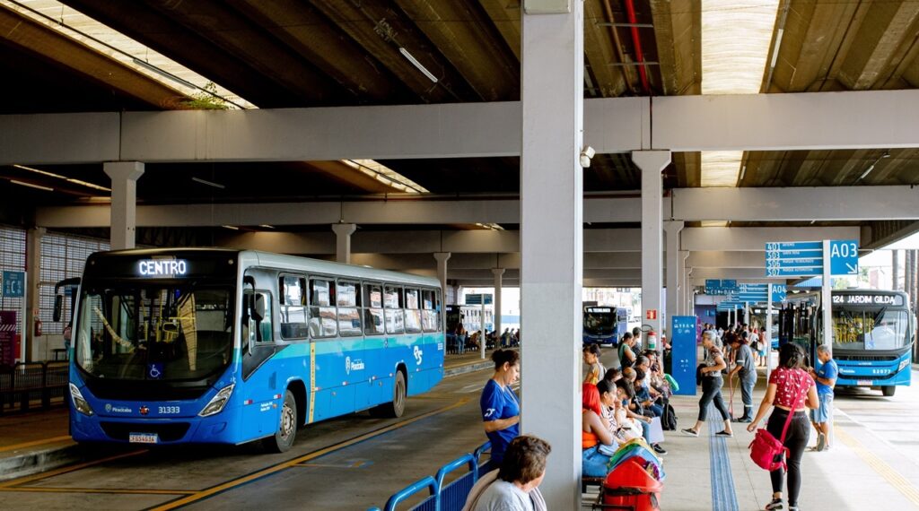 A imagem mostra um terminal de ônibus movimentado com vários ônibus azuis estacionados e pessoas esperando. Um ônibus azul com a palavra "CENTRO" em destaque na frente está mais próximo do espectador, posicionado em um ponto de ônibus. Outro ônibus azul, com o número 124 e o destino "JARDIM GILDA", está parado mais adiante. Há placas de sinalização com números de rota e destinos, como "A03" e "A04", suspensas acima dos pontos de ônibus. Pessoas estão sentadas em bancos, em pé ou caminhando pelo terminal, aguardando seus ônibus. A estrutura do terminal é caracterizada por pilares de concreto e um teto com iluminação. A luz do sol entra pelas aberturas do teto, criando um jogo de luz e sombra.