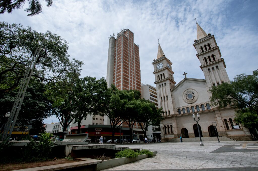 A imagem exibe uma praça com uma igreja imponente ao fundo, apresentando uma arquitetura clássica com duas torres de sino e uma fachada em tons claros. Uma das torres possui um relógio visível. Ao lado da igreja, ergue-se um edifício moderno e alto, com uma fachada em tijolos aparentes e detalhes em concreto.A praça em primeiro plano é pavimentada com pedras de diferentes tons e possui algumas áreas ajardinadas com árvores frondosas que criam sombra. Há também elementos de mobiliário urbano, como bancos e postes de iluminação. O céu está parcialmente nublado, com nuvens brancas e espalhadas em um fundo azul. Algumas pessoas circulam pela praça e pela calçada em frente aos estabelecimentos comerciais localizados na base de outros edifícios ao fundo, indicando um ambiente urbano movimentado.