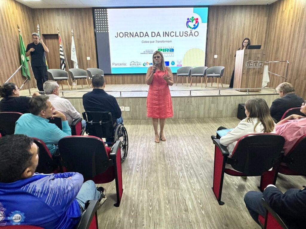 A imagem retrata um evento chamado "Jornada da Inclusão", com o subtítulo "Cotas que Transformam". No palco, uma mulher com um vestido coral está falando em um microfone, enquanto outra mulher está em pé atrás de um púlpito à direita. Há um telão ao fundo com o título do evento e logotipos. A plateia está sentada em cadeiras dispostas em fileiras. Destacam-se algumas pessoas com deficiência, como um homem em uma cadeira de rodas e outro que parece estar usando um aparelho auditivo. O ambiente parece ser uma sala de conferências ou auditório, com piso de madeira e paredes revestidas. Há bandeiras à esquerda do palco.
