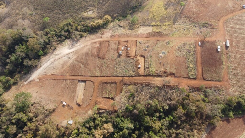 A imagem aérea revela um canteiro de obras em um terreno rural, com sinais de movimentação de terra e construção. No centro da imagem, observa-se um conjunto de lotes recém-esculpidos no solo, com algumas máquinas de construção, como retroescavadeiras, em operação. Uma estrada de terra batida serpenteia pelo local, conectando as diferentes áreas de trabalho. Ao redor, a vegetação nativa, composta por árvores e arbustos, contrasta com as áreas de terra exposta. Em uma das extremidades, um pequeno abrigo com cobertura e cercado sugere uma área de descanso ou armazenamento. Veículos, incluindo carros e caminhões, estão estacionados em pontos estratégicos do canteiro, indicando a atividade em andamento. No fundo, o terreno se estende por colinas com vegetação mais rasteira e áreas de cultivo.