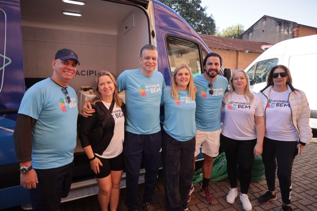 A imagem mostra um grupo de seis pessoas em frente a um veículo. Todos parecem estar vestindo camisetas azuis claras com um logotipo em cores vibrantes que diz "MOVIMENTO DO BEM". Da esquerda para a direita, vemos: 1. Um homem de boné azul, camiseta azul clara e bermuda azul escura. Ele está sorrindo para a câmera e tem óculos escuros pendurados na gola da camiseta. 2. Uma mulher com cabelo loiro, usando uma jaqueta preta sobre uma camiseta branca com o mesmo logotipo "MOVIMENTO DO BEM". Ela está sorrindo e tem um braço ao redor do ombro do homem ao seu lado. 3. Um homem sorrindo, vestindo uma camiseta azul clara e calça de moletom azul escura. 4. Uma mulher loira, sorrindo, vestindo uma camiseta azul clara com o logotipo. 5. Um homem com barba, vestindo uma camiseta azul clara com o logotipo e bermuda branca. Ele também está sorrindo. 6. Uma mulher com cabelo escuro, usando uma jaqueta branca acolchoada sobre uma camiseta branca com o logotipo "MOVIMENTO DO BEM". Ela está usando óculos escuros e sorrindo levemente. O veículo atrás deles parece ser uma van ou um pequeno ônibus, com a porta traseira aberta. Há também uma parte branca de outro veículo visível à direita. O fundo sugere um ambiente externo, com uma parede de tijolos e algumas árvores ao fundo. As pessoas parecem estar participando de um evento ou atividade relacionada ao "Movimento do Bem".