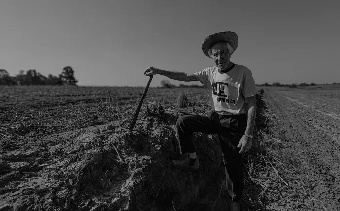 A imagem em preto e branco mostra um idoso sentado em uma elevação de terra em um campo. Ele usa um chapéu de palha e uma camiseta com um logo que diz "DC Sports Co.". Em sua mão direita, ele segura uma vara ou cajado, apoiando-se nele. O campo ao redor parece ter sido cultivado, com sulcos visíveis no solo e restos de vegetação. Ao fundo, o céu está claro e uma linha de árvores pode ser vista no horizonte. A iluminação sugere que a foto foi tirada durante o dia, com o sol iluminando a cena. A composição da imagem foca no idoso e em sua conexão com a terra, transmitindo uma sensação de trabalho árduo e experiência.