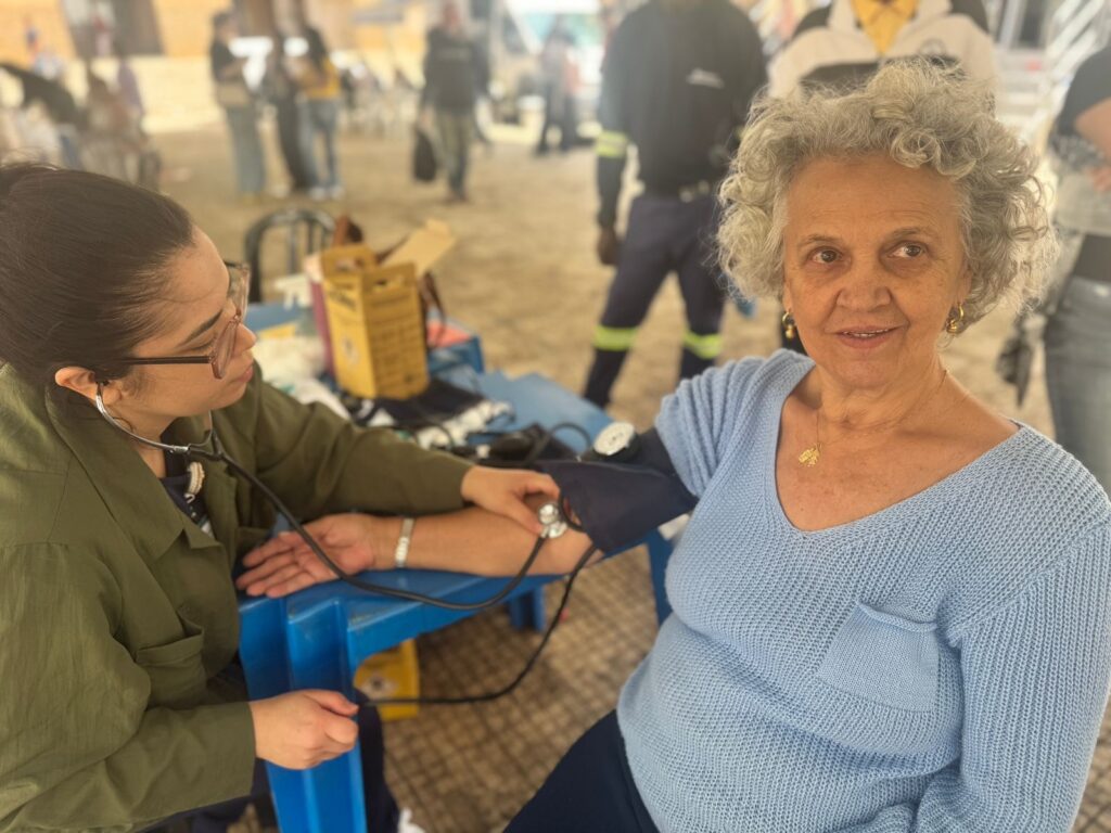 A enfermeira, usando óculos e jaleco verde, está em primeiro plano à esquerda, com um estetoscópio em uso. Ela está sentada em uma mesa azul e executa o exame de pressão arterial em uma paciente idosa. A paciente, com cabelo grisalho e encaracolado, usa um suéter azul claro e sorri para a câmera. Ao fundo, pessoas e algumas caixas de suprimentos médicos compõem o cenário, que parece ser um evento ao ar livre.