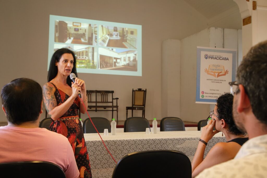 A mulher de cabelos pretos está em pé, segurando um microfone e falando para uma audiência. Ela usa um vestido estampado com cores vibrantes. Atrás dela, um projetor exibe uma grade de quatro imagens mostrando o interior e o exterior de edifícios. Há uma mesa com garrafas de água e copos, e cadeiras vazias. Ao lado, uma faixa azul com o logotipo da "Escola Legislativo" de Piracicaba e informações de contato. No primeiro plano, as costas de duas pessoas na audiência são visíveis.