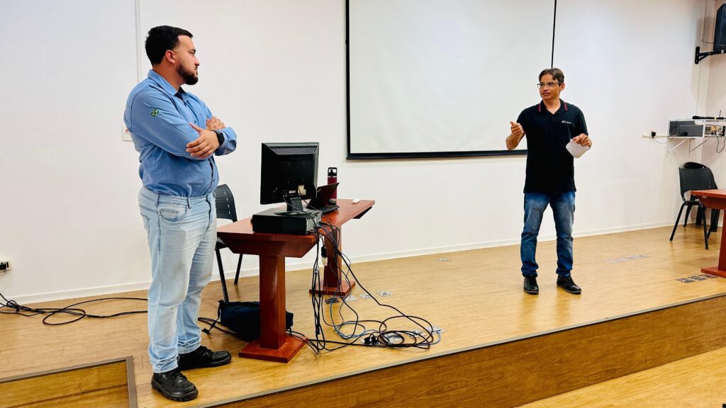 A imagem mostra duas pessoas em uma sala de apresentação. A pessoa à esquerda está de pé, com os braços cruzados, vestindo uma camisa azul e calças jeans claras. Ela tem barba e cabelo escuro. A pessoa à direita está em pé, de frente para a tela de projeção, vestindo uma camisa preta e calças jeans. Ela está gesticulando com as mãos e segurando um papel. Há uma mesa com um computador e outros equipamentos no centro da sala, e uma tela de projeção em branco ao fundo. O piso é de madeira clara, e há cadeiras nas laterais.