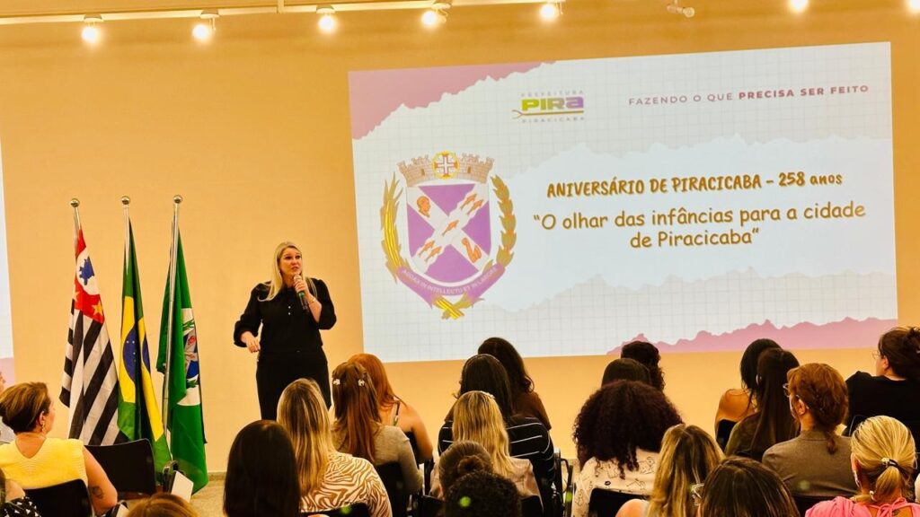A imagem mostra uma mulher loira, vestida de preto, falando em um microfone enquanto está em pé em frente a uma audiência. Há três bandeiras atrás dela: a bandeira do Estado de São Paulo, a bandeira do Brasil e a bandeira do município de Piracicaba. Ao fundo, um telão exibe o brasão de Piracicaba, o logotipo da prefeitura e a frase "Aniversário de Piracicaba - 258 anos" e "O olhar das infâncias para a cidade de Piracicaba". A plateia, composta principalmente por mulheres, está sentada e voltada para a palestrante. O ambiente parece ser uma sala de eventos ou auditório.