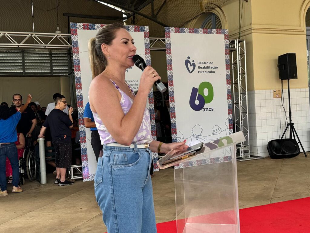 A mulher loira está falando em um microfone e segurando um tablet. Ela está usando um top sem mangas com estampa floral e calça jeans. Ela está em frente a um pódio transparente. Atrás dela, há um banner com o logotipo do "Centro de Reabilitação Piracicaba" e o número "60 anos". Há pessoas ao fundo, incluindo uma em cadeira de rodas.