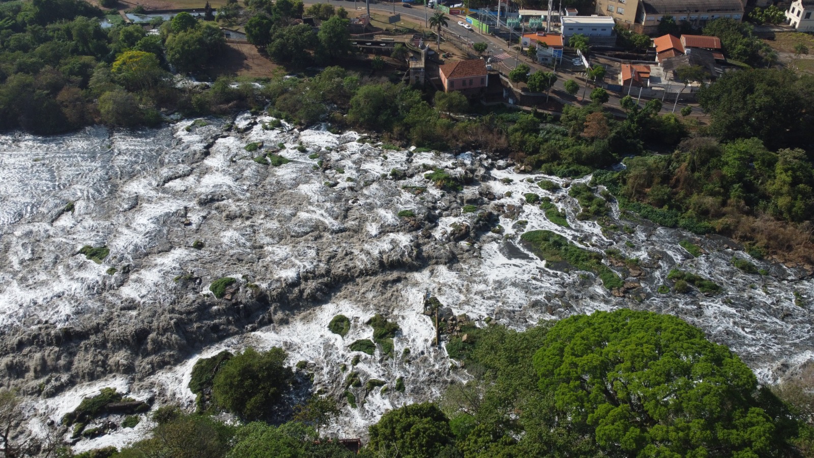 A imagem mostra um rio com águas turbulentas e espumosas, com pedras e vegetação emergindo da água. Na margem direita, observa-se uma área urbana com casas, edifícios e uma estrada com carros. A vegetação é densa, com árvores altas e arbustos ao longo da margem do rio e nas áreas mais afastadas. O céu está claro, sugerindo um dia ensolarado. A perspectiva é de um ponto elevado, possivelmente de um drone ou avião, que oferece uma visão panorâmica da paisagem.