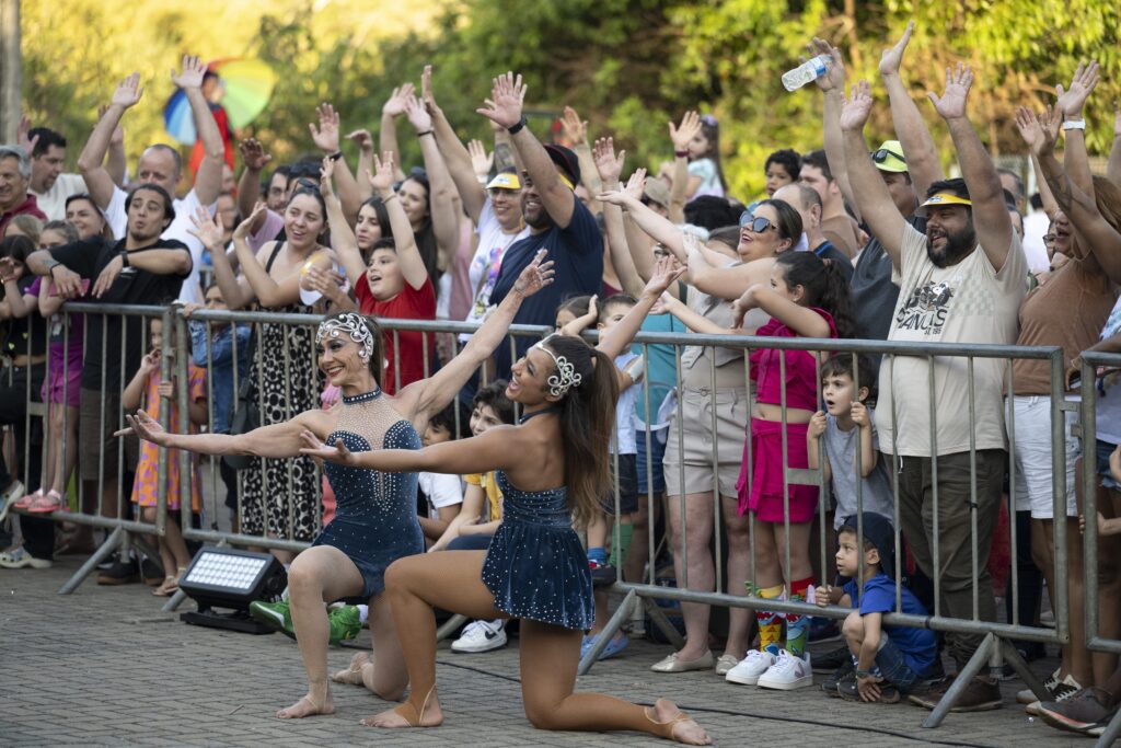 Duas artistas em trajes de apresentação azul com brilho posam sorrindo à frente do público, que levanta os braços animado atrás das grades de proteção.