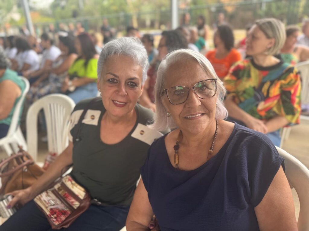 A imagem mostra duas mulheres mais velhas sentadas em cadeiras brancas, sorrindo para a câmera. Elas parecem estar em um evento ao ar livre, com outras pessoas sentadas em cadeiras desfocadas ao fundo.A mulher à esquerda tem cabelos grisalhos curtos e usa uma blusa verde escura com detalhes em branco nos ombros. Ela tem um sorriso radiante e usa maquiagem leve. Uma bolsa com estampa floral está apoiada em seu colo.A mulher à direita tem cabelos grisalhos mais longos e usa óculos de aro grosso. Ela veste uma blusa azul marinho e um colar com contas de madeira. Seu sorriso é mais contido, mas ela também parece feliz.O fundo da imagem está desfocado, mas é possível ver várias outras pessoas sentadas, sugerindo que o evento pode ser uma reunião comunitária, um culto religioso ou uma apresentação. O ambiente parece ser diurno e ensolarado.