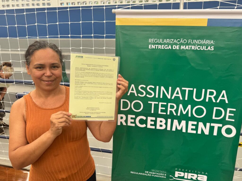 A imagem mostra uma mulher sorridente em ambiente interno, segurando um documento na mão direita, ao lado de um banner verde com texto em branco e azul-claro. O banner anuncia a “Regularização Fundiária” e a “Entrega de Matrículas”, indicando que o evento está relacionado à formalização de propriedades ou terrenos. Também é mencionada a “Assinatura do Termo de Recebimento”, promovida pela Secretaria de Habitação e Regularização Fundiária da Prefeitura de Piracicaba.