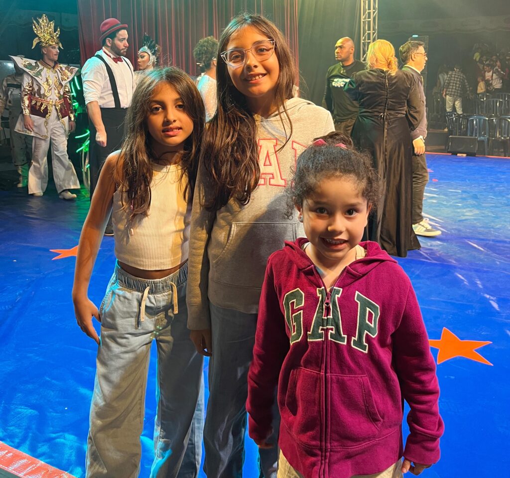 Três meninas sorridentes posam em frente ao palco do circo, duas de cabelo solto e uma de moletom rosa.