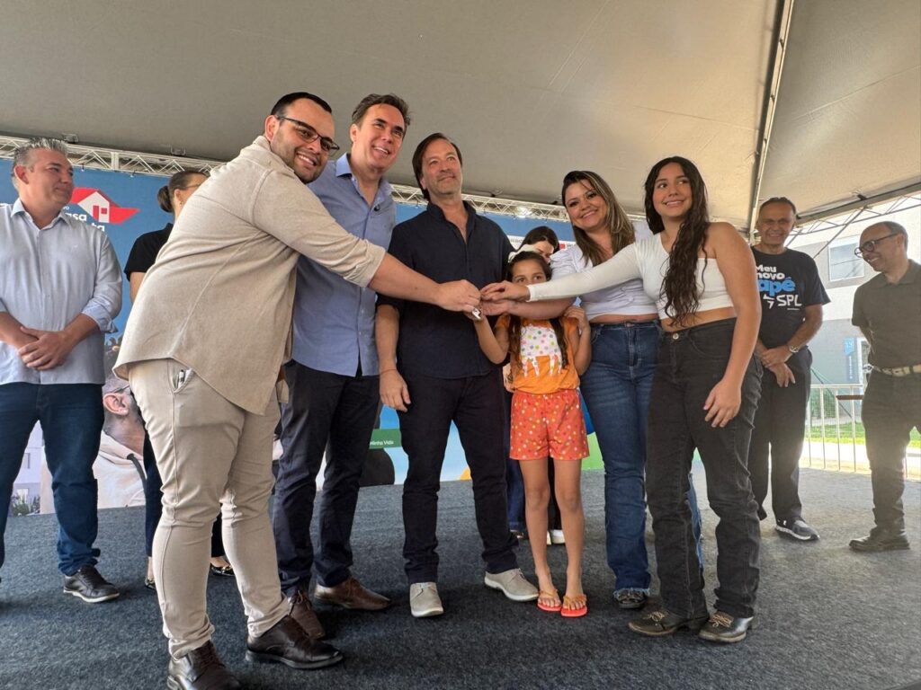 A imagem mostra um grupo de pessoas em um palco, possivelmente durante um evento. Há seis pessoas visíveis em primeiro plano. À esquerda, um homem de camisa clara e calça jeans está de pé, com as mãos cruzadas. Ao lado dele, um homem com óculos, camisa bege e calça cáqui está curvado, sorrindo e abraçando o homem ao seu lado. Este último, vestindo uma camisa azul e calça preta, também sorri. Mais à direita, um homem com camisa escura e calça preta está em pé. No centro, uma menina com uma blusa laranja estampada e shorts laranjas está entre os adultos, com as mãos dadas com uma mulher e um homem. A mulher, ao lado da menina, usa uma blusa branca e calça jeans. Ao lado dela, uma jovem com uma blusa branca curta e calça jeans preta também está presente. Ao fundo, há um banner com um logo de casa azul e vermelho e texto em branco. O palco parece estar ao ar livre, com uma estrutura de teto visível acima.