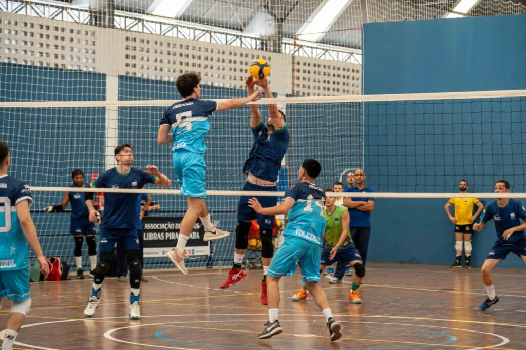 A imagem mostra uma partida de vôlei em uma quadra interna. Dois jogadores estão em primeiro plano, um saltando para bloquear uma bola de vôlei amarela e azul, enquanto o outro se prepara para recebê-la. Ambos os jogadores usam uniformes de vôlei, um azul escuro com detalhes em azul claro e o outro em azul claro com detalhes em azul escuro. A rede de vôlei está esticada entre eles. Ao fundo, outros jogadores e espectadores podem ser vistos. A quadra tem um piso de madeira polida e as paredes são azuis. Uma rede de proteção preta cobre a parte superior da quadra. Há uma placa visível na parede atrás dos jogadores, com texto em português, parte dele ilegível. A iluminação parece ser artificial, vinda de cima. A cena captura um momento dinâmico de ação no jogo de vôlei.