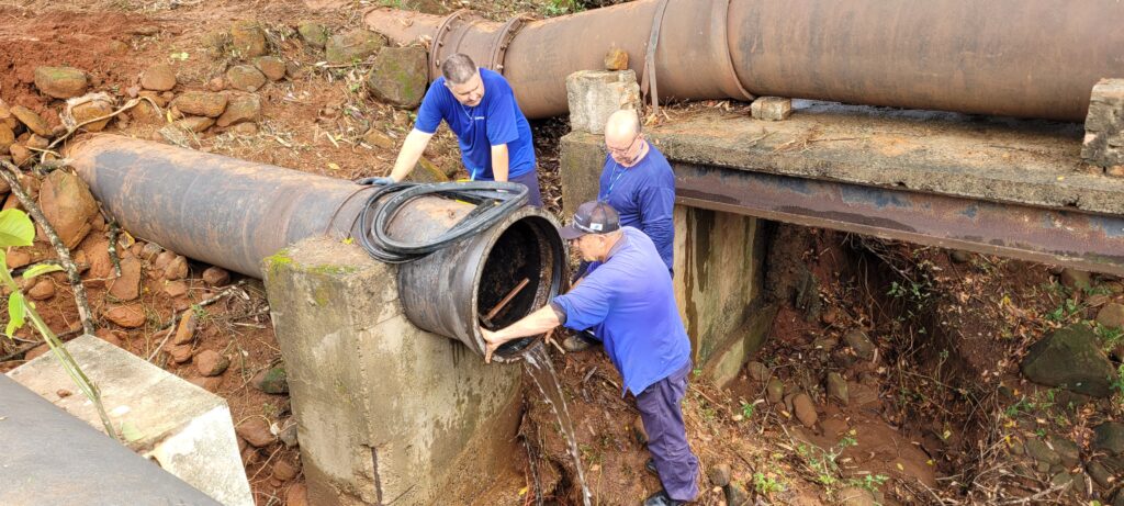 Três homens em camisetas azuis estão trabalhando em um grande cano de água. Um homem está curvado sobre um cano menor que está saindo de um cano maior. Ele está segurando uma peça de borracha em forma de anel e parece estar trabalhando na junta. Um segundo homem, com óculos, está em pé atrás dele, observando o trabalho. Um terceiro homem está mais atrás, com as mãos apoiadas no cano maior. O cano maior está apoiado em uma estrutura de concreto e metal, e parece estar em um ambiente externo com terra e pedras ao redor. Há água saindo do cano menor.