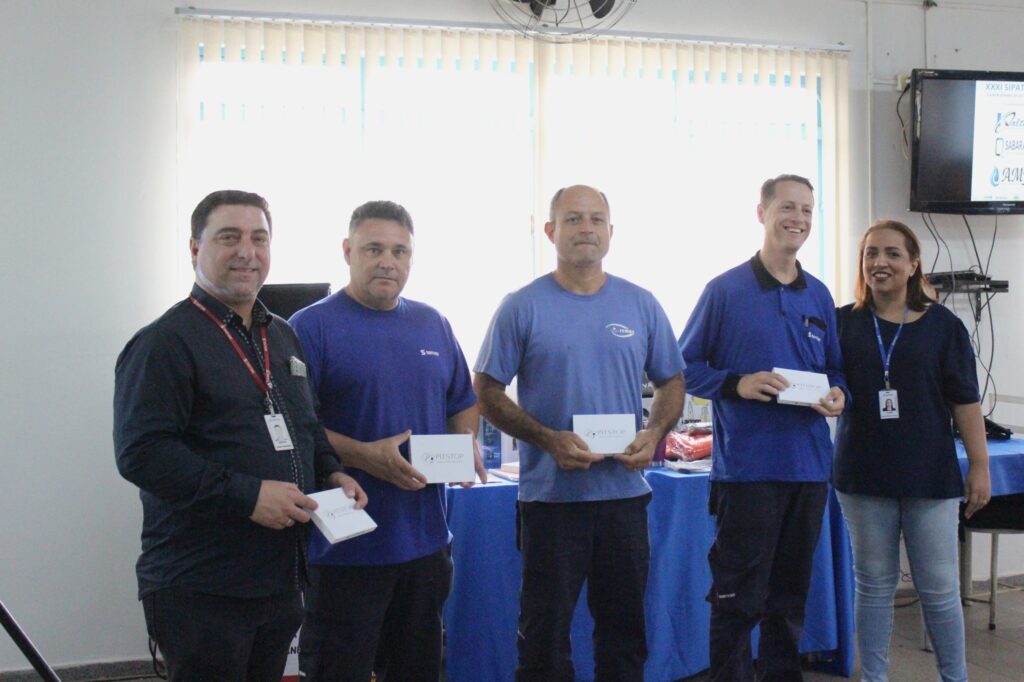A imagem mostra cinco pessoas em pé, em frente a uma parede branca com cortinas. Quatro homens e uma mulher estão alinhados, segurando pequenos pacotes brancos com o logotipo "PIT STOP". O homem mais à esquerda, vestindo uma camisa preta e um crachá vermelho, sorri para a câmera. Ao seu lado, um homem em uma camiseta azul também sorri. O terceiro homem da esquerda para a direita, vestindo uma camiseta azul clara, segura um pacote e olha para a frente. O quarto homem, em uma camiseta azul escura, sorri e segura um pacote. A mulher à direita, vestindo uma blusa azul escura e jeans, também sorri e usa um crachá. Ao fundo, uma televisão exibe o texto "XXI SIPAT" e outros logotipos, sugerindo que o evento é uma palestra ou celebração relacionada à segurança no trabalho. Mesas cobertas com toalhas azuis estão dispostas atrás das pessoas.