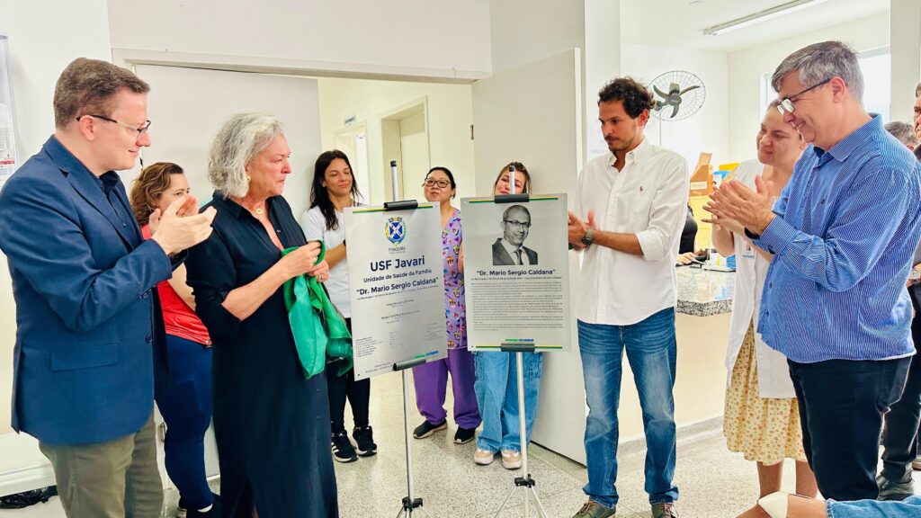 A imagem captura um momento de reconhecimento e celebração na USF Javari, uma Unidade de Saúde da Família. Várias pessoas estão reunidas, algumas aplaudindo, em um ambiente interno, possivelmente um corredor ou área de entrada da unidade de saúde. Em primeiro plano, uma mulher vestindo um vestido preto está segurando um lenço ou faixa verde. À sua esquerda, um homem em um terno azul e óculos também está aplaudindo, olhando para a mulher. Atrás deles, outras pessoas são visíveis, algumas também aplaudindo. Dois grandes painéis de exibição estão em destaque. O painel à esquerda tem o texto "PRACCABA USF Javari Unidade de Saúde da Família 'Dr. Mario Sergio Caldana'". O painel à direita também exibe o nome "Dr. Mario Sergio Caldana" junto com um retrato de um homem usando óculos e terno. A atmosfera geral sugere um evento em homenagem ao Dr. Mario Sergio Caldana e à USF Javari.