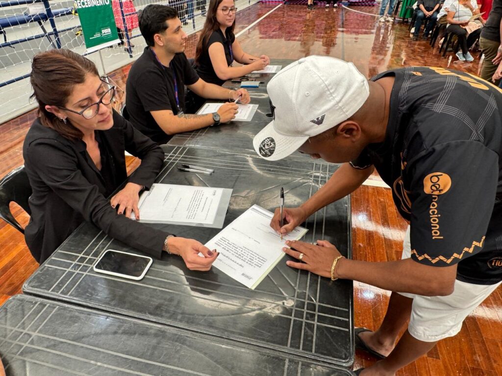 A imagem retrata um momento de atendimento administrativo em um ginásio coberto, com piso de madeira polida e uma parede com rede ao fundo. Algumas pessoas estão sentadas à mesa, auxiliando outras que estão em pé e preenchendo formulários. Sobre a mesa de superfície marmorizada, há documentos, canetas e um celular, indicando um processo formal de registro ou declaração. Em destaque, uma mulher de óculos, vestida de preto, orienta alguém que assina uma “Declaração de Residência”.