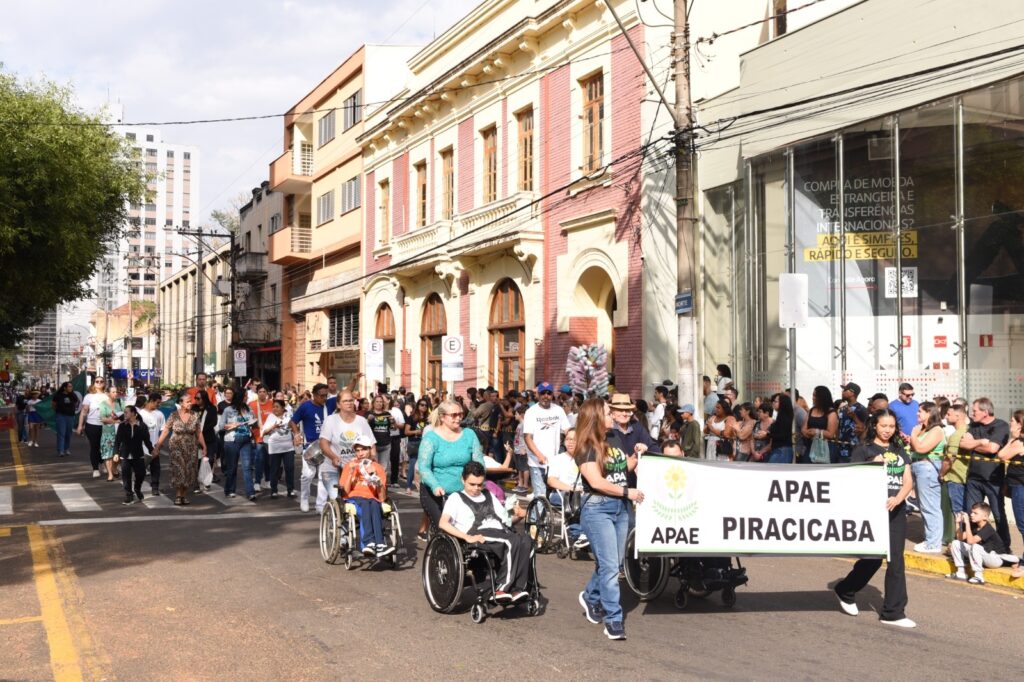 A imagem mostra um desfile de rua com muitas pessoas. No centro do desfile, há um grupo de pessoas carregando uma faixa com os dizeres "APAE PIRACICABA". Algumas pessoas estão em cadeiras de rodas, participando do desfile. Ao fundo, há edifícios com arquitetura clássica e moderna, e um céu parcialmente nublado. A rua está decorada com faixas e balões. A atmosfera geral parece ser de celebração e inclusão.