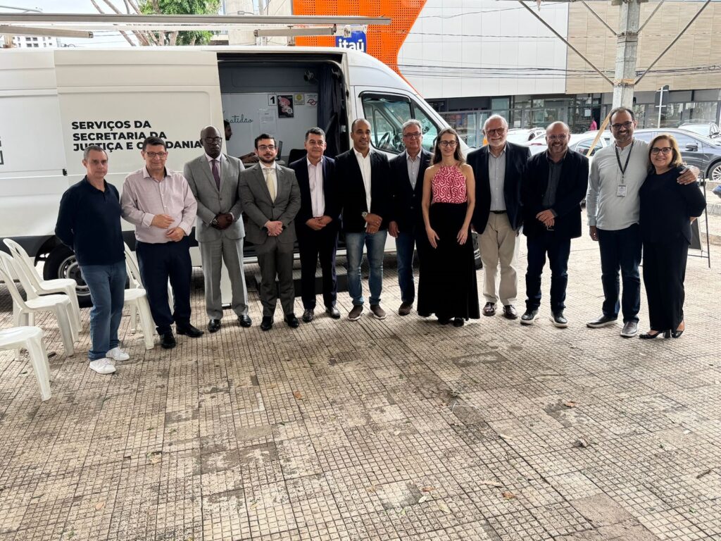 A fotografia mostra um grupo de onze pessoas em frente a uma van branca com o texto "SERVIÇOS DA SECRETARIA DA JUSTIÇA E CIDADANIA" escrito em preto. O grupo é composto por seis homens e cinco mulheres. Todos estão de pé e posando para a câmera. A van parece ser um veículo móvel de prestação de serviços. A maioria das pessoas está vestida formalmente, com ternos e vestidos. O fundo da imagem é composto por um edifício e algumas árvores. A iluminação é natural, sugerindo que a foto foi tirada durante o dia. A textura do chão, feito de ladrilhos quadrados em tons de cinza e bege, é evidente na parte inferior da imagem.