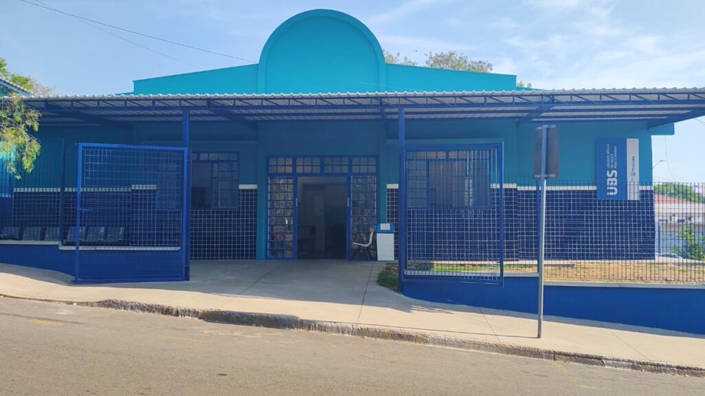 A imagem mostra a fachada de uma Unidade Básica de Saúde (UBS) em um dia ensolarado. O edifício é predominantemente pintado em tons de azul claro e azul escuro. A parte superior da estrutura é um azul mais vibrante, com uma marquise metálica azul cobrindo a entrada principal e as janelas laterais. A marquise é sustentada por colunas azuis. A entrada principal é composta por portas de vidro com caixilhos azuis, permitindo vislumbrar o interior do prédio, onde há algumas cadeiras e uma lixeira. À esquerda e à direita da entrada, há janelas grandes, também com caixilhos azuis. A parte inferior da fachada é revestida com azulejos em um tom de azul escuro, formando um padrão quadriculado. Grades azuis de metal protegem algumas áreas, como a entrada lateral e uma área de espera externa com bancos. Na lateral direita da edificação, há um poste com uma placa vertical que exibe o logo "UBS" e informações textuais, como "UNIDADE BÁSICA DE SAÚDE DE SAO JOAQUIM" e o nome "Pescaria". O edifício está localizado em uma rua com uma leve inclinação, e a calçada em frente à UBS é de concreto. Ao fundo, é possível ver algumas árvores e construções. O céu está limpo e azul, indicando um dia claro.