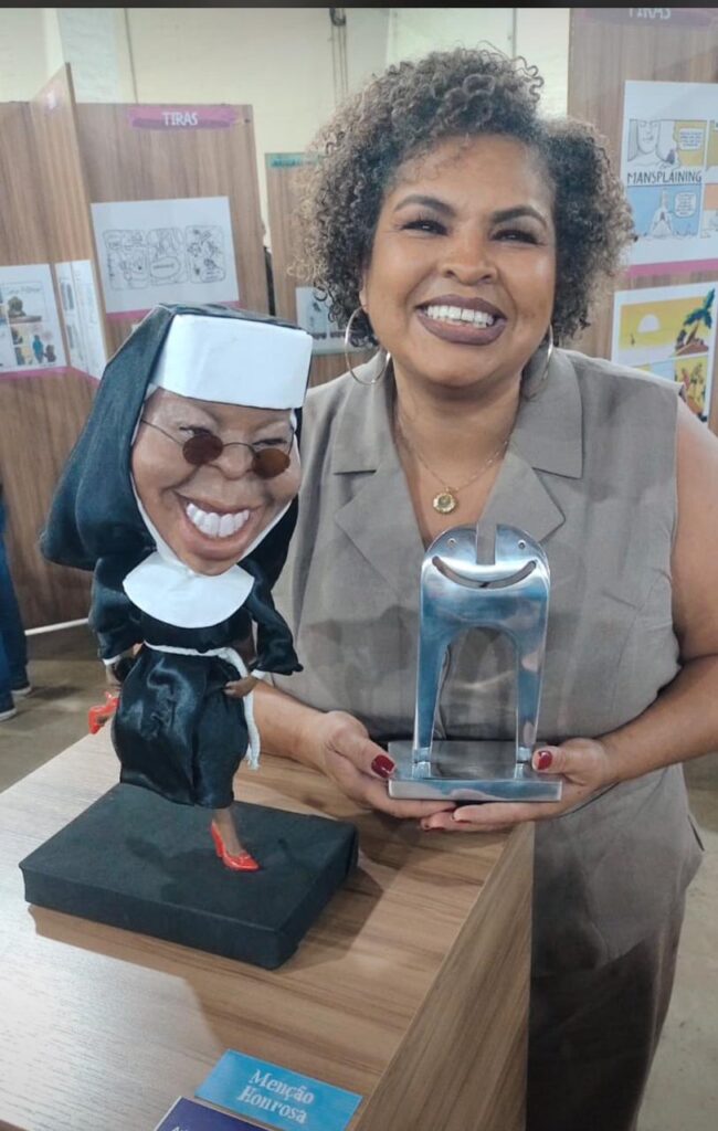 A mulher na foto parece estar feliz e orgulhosa, segurando um troféu e uma caricatura. A caricatura é de uma freira, com um sorriso largo e óculos escuros. Ela está vestida com um hábito de freira preto e branco, e usa sapatos vermelhos. O troféu é feito de metal prateado e tem um design abstrato, com um rosto sorridente no topo. Ao fundo, há um painel com várias tiras de quadrinhos. Uma delas é intitulada "MANSPLAINING", e outra parece ser sobre tiras de jornal. Há também uma placa azul na frente da mesa que diz "Menção Honrosa". A mulher tem cabelo cacheado escuro e usa um colar com um pingente dourado. Suas unhas estão pintadas de vermelho. A iluminação na foto é suave, e o fundo é um pouco desfocado, o que ajuda a destacar a mulher e os objetos que ela está segurando.