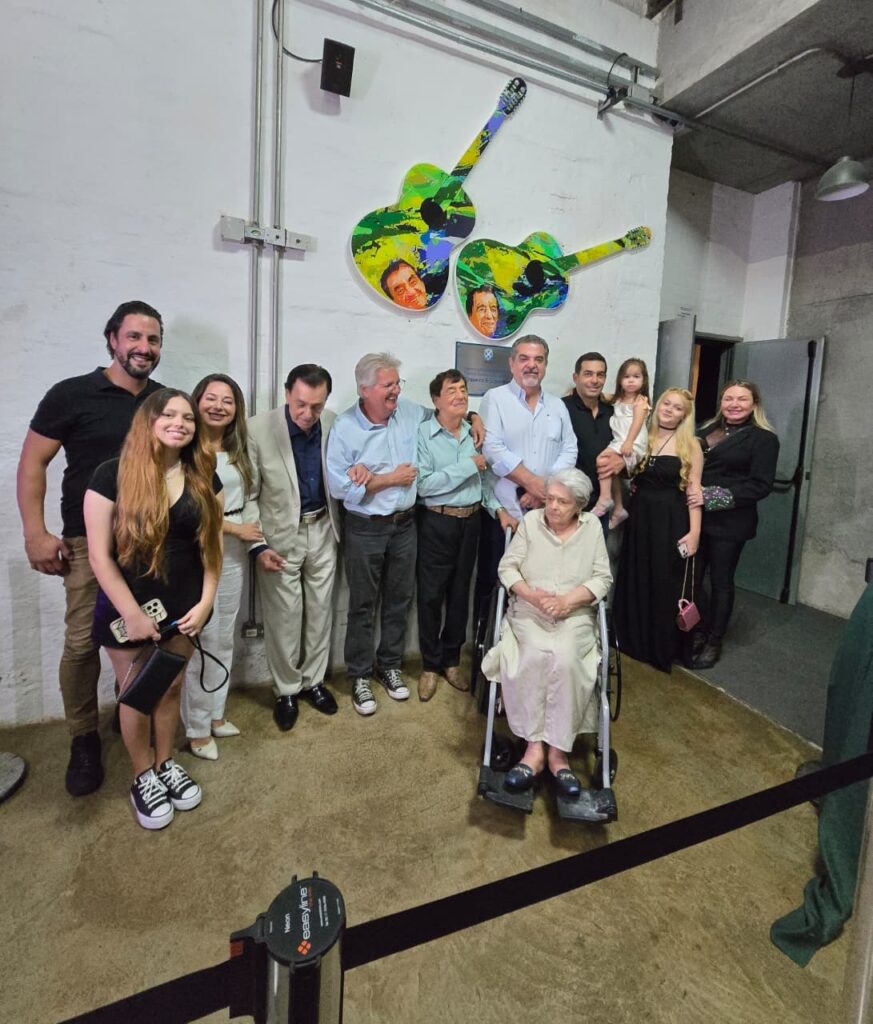 Na imagem aparecem homens e mulheres em enfileirados e a frente uma senhora em cadeiras de rodas. Estão posicionados em uma entrada de um teatro. Na parede aparece o desenho de duas violas de cor verde com detalhes em preto, estampada com um rosto de dois homens.
