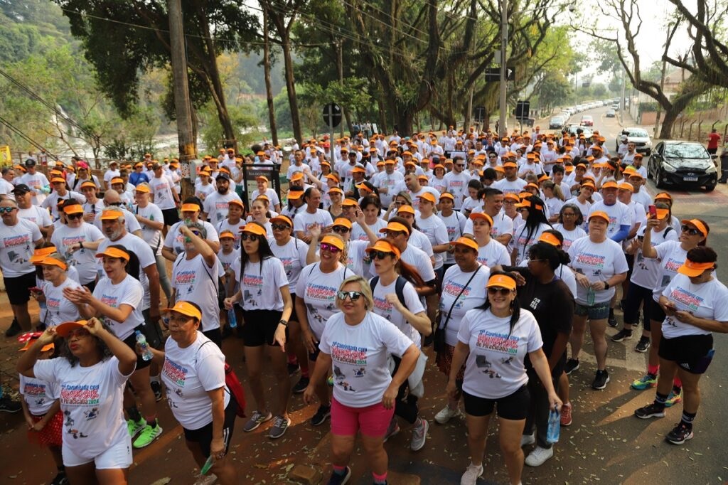 O evento retratado na imagem é uma caminhada em grupo, com a maioria dos participantes vestindo camisetas brancas e bonés laranja. A cena se passa em uma rua arborizada, com carros e edifícios ao fundo, sugerindo um ambiente urbano. A atmosfera parece ser de confraternização e atividade física ao ar livre, com pessoas de diversas idades presentes.