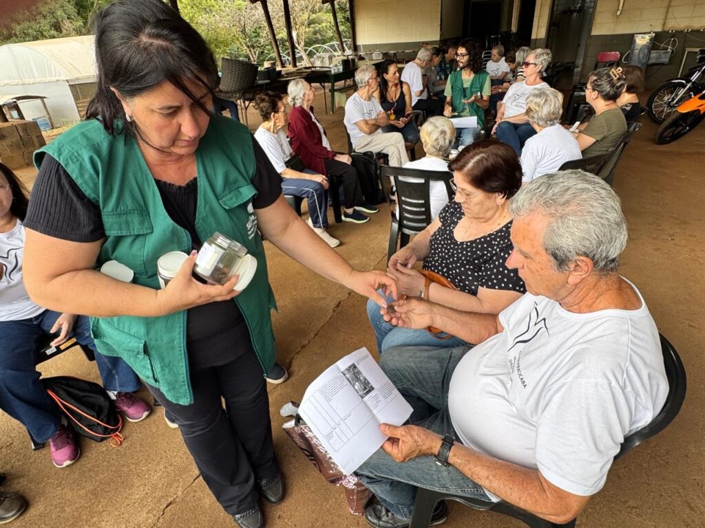A mulher de colete verde está mostrando algo em um pote de vidro para um homem sentado. Ele está segurando um papel com informações. Ao redor deles, há um grupo de pessoas sentadas, ouvindo ou observando a demonstração. O ambiente parece ser ao ar livre, possivelmente em um evento ou workshop.