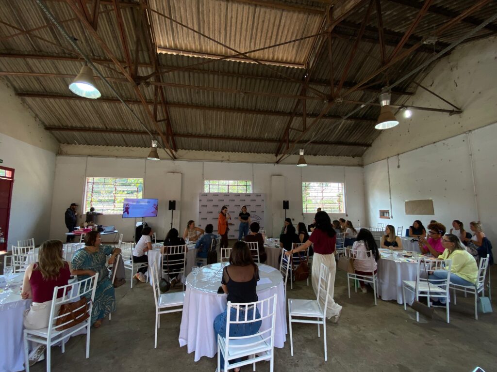 A imagem mostra um evento ou workshop acontecendo em um espaço amplo com um telhado de zinco aparente e estruturas metálicas. Há várias mesas redondas cobertas com toalhas brancas, dispostas pelo local, com cadeiras brancas ao redor. No centro do espaço, há um palco improvisado com um telão exibindo uma apresentação, duas mulheres em pé falando para a audiência e um banner ao fundo com logotipos. A audiência, composta majoritariamente por mulheres, está sentada às mesas, atenta à apresentação. Algumas pessoas estão em pé, conversando ou se movimentando. A iluminação vem de lâmpadas penduradas no telhado e da luz natural que entra por algumas janelas.