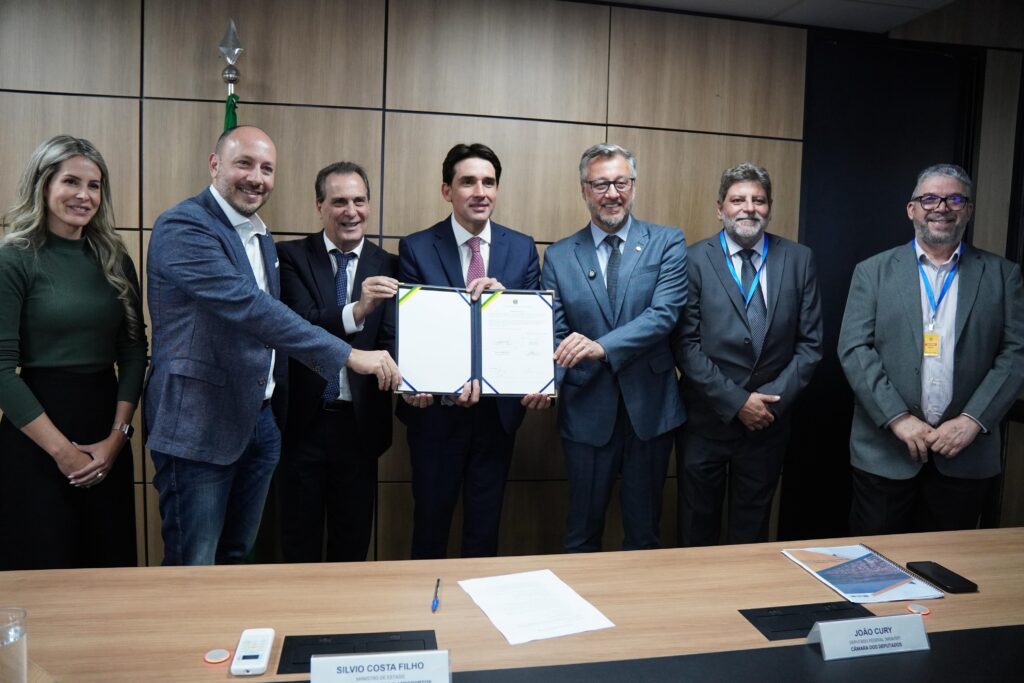 A imagem mostra um grupo de sete pessoas, seis homens e uma mulher, em pé atrás de uma mesa. Eles estão em um ambiente interno, possivelmente um escritório ou sala de reuniões, com paredes de madeira ao fundo. No centro da imagem, dois homens estão segurando um documento aberto, que parece ser um contrato ou um termo de acordo. Ambos sorriem para a câmera. Os outros homens e a mulher também estão sorrindo e olhando para a frente. A mulher à esquerda está vestindo uma blusa verde escura e uma saia preta. O homem ao lado dela usa um blazer azul escuro sobre uma camisa branca e jeans. Os outros homens vestem ternos e gravatas, alguns com crachás pendurados no pescoço. Na mesa, há alguns objetos, incluindo um celular, um copo com água e alguns papéis, um deles com o nome "SILVIO COSTA FILHO MINISTRO DE ESTADO MINISTÉRIO DOS PORTOS E AEROPORTOS" e outro com "JOÃO CURY DEPUTADO FEDERAL (PSDB-SP CÂMARA DOS DEPUTADOS". A iluminação é clara e a foto parece ter sido tirada em um evento oficial ou uma cerimônia de assinatura.