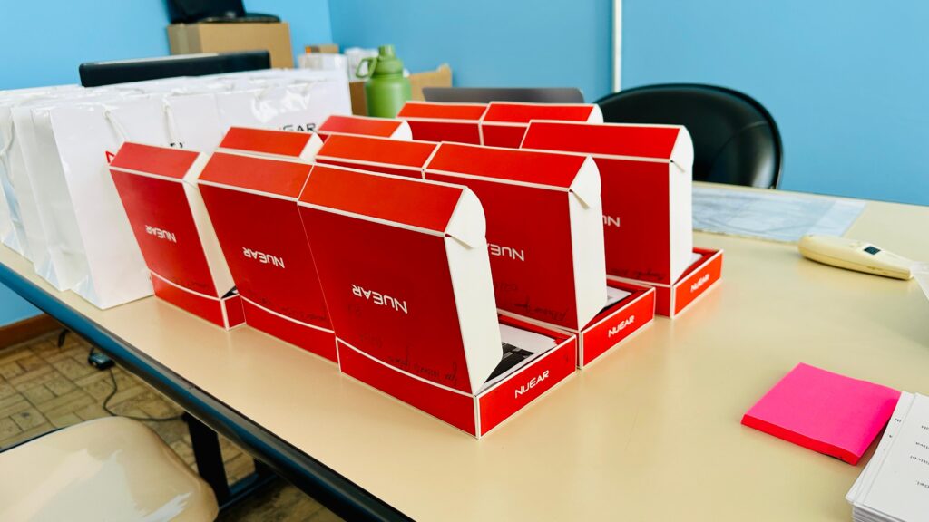 A imagem mostra uma mesa de escritório com vários itens dispostos sobre ela.

No lado esquerdo, há várias sacolas brancas de papel, algumas com o nome "NUAR" ou "CAR" visível.

À frente das sacolas, há uma linha de caixas vermelhas com detalhes brancos, cada uma com a marca "NUAR" impressa. As caixas parecem ser de produtos eletrônicos ou similares, pois algumas estão abertas, revelando o conteúdo interno.

No lado direito da mesa, há um objeto branco que se assemelha a um controle remoto ou dispositivo eletrônico, e um pequeno bloco de notas rosa brilhante. Há também uma pilha de papéis ao lado do bloco de notas.

Ao fundo, pode-se ver uma cadeira de escritório preta e uma parede azul. Há também uma garrafa verde e caixas de papelão em segundo plano, sugerindo um ambiente de escritório ou estoque.