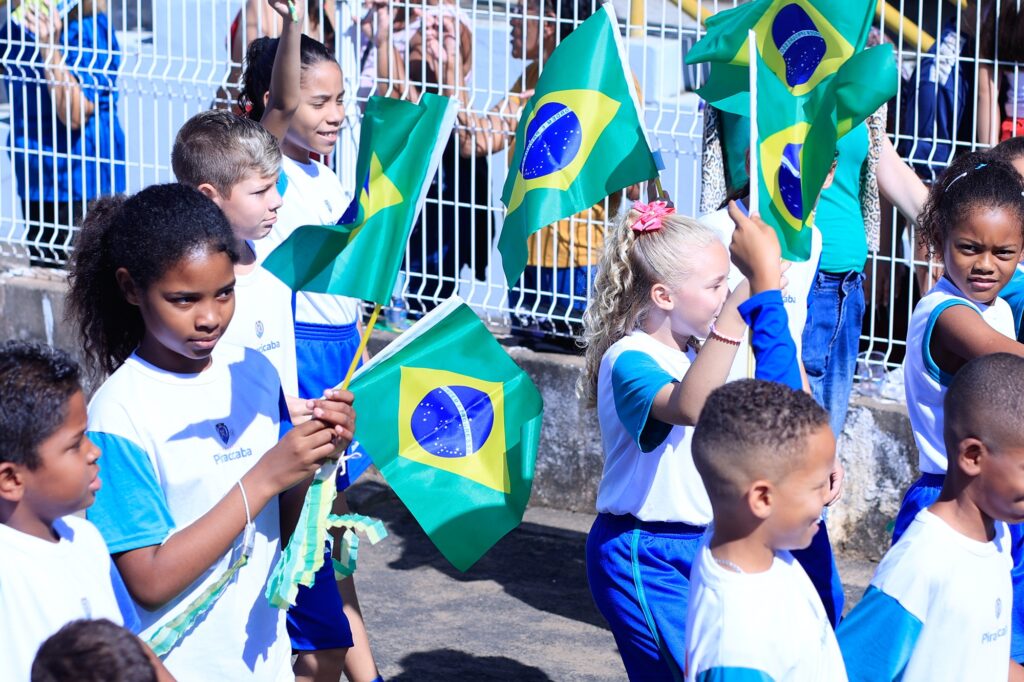 A imagem retrata um grupo de crianças participando de um desfile ou evento público, vestidas com uniformes que combinam camisetas brancas com mangas azuis e calças azuis. Elas caminham alegremente por uma rua ensolarada, muitas segurando bandeiras do Brasil, o que sugere um momento de celebração nacional ou cívica.