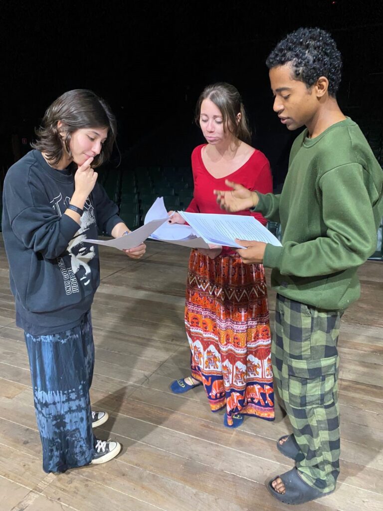 Três atores da Cia. Ceta em pé no palco, concentrados, segurando folhas com o roteiro da peça E Amanhã?. Eles discutem o texto, vestidos de forma descontraída, em roupas largas e coloridas.