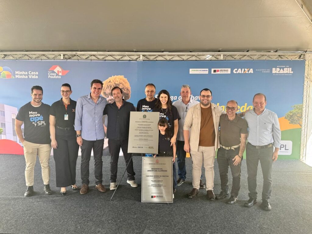 A imagem mostra um grupo de pessoas em frente a um banner com logotipos de programas habitacionais e de governo. As pessoas parecem estar em um evento de inauguração ou lançamento de um empreendimento imobiliário. No centro, há uma placa com o título "RESIDENCIAL JARDIM DAS ALAMEDAS" e nomes de pessoas, possivelmente autoridades ou responsáveis pelo projeto. Há também logotipos de "Minha Casa Minha Vida", "Casa Paulista", "CAIXA", "Governo do Brasil" e "Governo de São Paulo", indicando uma parceria público-privada para a construção de moradias. As pessoas no grupo incluem homens e mulheres de diferentes idades, vestindo roupas casuais e formais. Alguns seguram a placa, enquanto outros posam para a foto. O ambiente sugere um dia ensolarado, com uma estrutura de palco ou cobertura ao fundo.