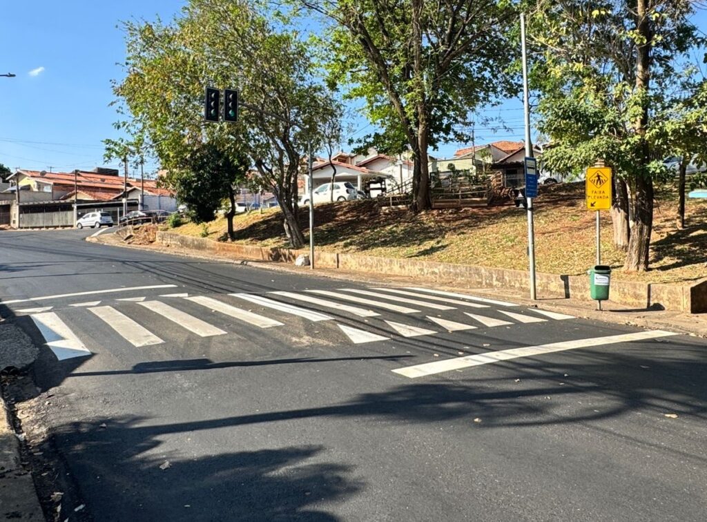 A imagem mostra uma rua com uma faixa de pedestres recém-pintada. O asfalto ao redor da faixa de pedestres parece novo, com linhas brancas nítidas. Há um semáforo no lado esquerdo da imagem, com as luzes verde e amarela acesas.No lado direito da rua, há uma placa amarela com um símbolo de pedestre e a inscrição "FAIXA ELEVADA" em letras pretas. Ao lado da placa, há uma lixeira verde. A rua está ladeada por árvores frondosas e casas com telhados vermelhos ao fundo. O céu está azul e claro, indicando um dia ensolarado. As sombras das árvores projetam-se sobre a rua, criando um padrão interessante.