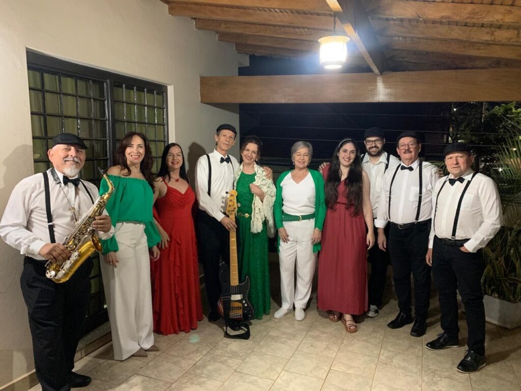 A imagem mostra um grupo de nove pessoas posando para uma foto. Eles parecem estar em um evento ao ar livre, possivelmente um show ou uma festa, com um telhado de madeira acima e iluminação ambiente. A maioria das pessoas está vestida de forma elegante, com vários homens usando suspensórios e gravatas borboleta, e as mulheres usando vestidos ou conjuntos de cores vibrantes. Dois membros do grupo estão segurando instrumentos musicais: um homem à esquerda está com um saxofone dourado, e uma mulher no centro está com uma guitarra elétrica verde. O grupo parece estar posicionado em um pátio ou varanda, com um piso de ladrilho visível. A iluminação sugere que a foto foi tirada à noite ou em um ambiente com pouca luz natural. As expressões faciais do grupo indicam que eles estão felizes e posando para a ocasião.