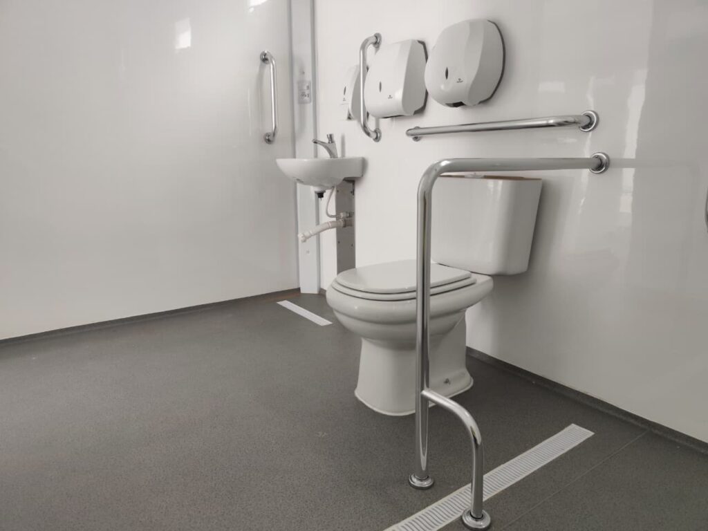 A imagem mostra um banheiro acessível, com paredes brancas e piso cinza texturizado. Há um vaso sanitário branco com assento e tampa, cercado por barras de apoio cromadas em diferentes posições para auxiliar na mobilidade. À esquerda do vaso sanitário, há uma pia pequena com torneira cromada e, acima dela, um dispensador de papel toalha e outro dispensador, possivelmente de sabão, montados na parede. Uma barra de apoio vertical cromada está fixada na parede ao lado da pia. Na parede atrás do vaso sanitário, há duas barras de apoio horizontais cromadas, uma mais alta e outra mais baixa, posicionadas estrategicamente para oferecer suporte. O piso é cinza e parece ser um material antiderrapante, com uma linha branca fina marcada no chão, possivelmente indicando uma rota ou área de segurança. Há também uma grade de drenagem linear discreta no piso, perto do vaso sanitário. A iluminação é clara e uniforme, destacando a limpeza e funcionalidade do espaço.