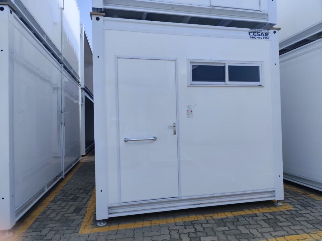 A imagem mostra uma unidade modular branca, provavelmente um banheiro ou escritório portátil, estacionada em um pátio de concreto com marcações amarelas no chão. A unidade tem uma porta branca com uma maçaneta prateada e uma barra de apoio. Acima da porta, há uma janela com duas divisões. No canto superior direito da unidade, há um adesivo com o logotipo "CESAR" e o número de telefone "0800 945 9500". Ao lado da unidade, empilhadas em primeiro plano, há outras unidades modulares brancas.