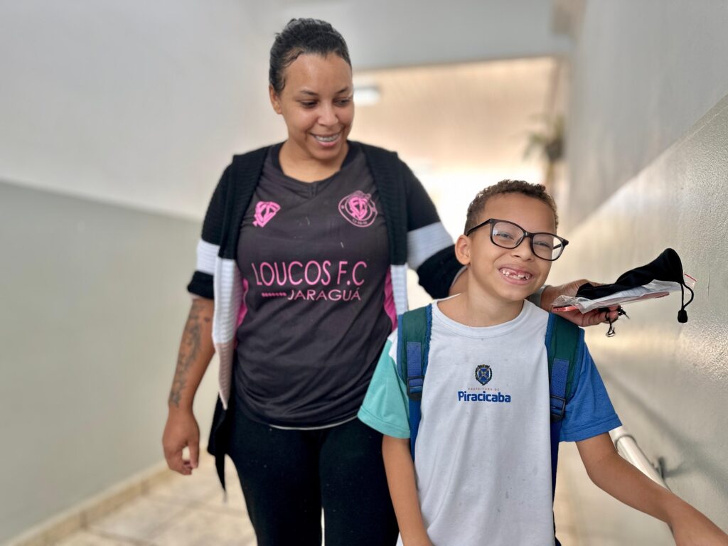 A imagem mostra uma mulher e um menino em um corredor. A mulher, com o cabelo preso em um coque, veste uma camiseta preta com detalhes em rosa e um cardigã preto e branco. Ela tem uma tatuagem no braço esquerdo e usa um aparelho nos dentes. O menino, que usa óculos, está à frente dela e veste uma camiseta branca com detalhes em azul e verde, além de uma mochila. Ele sorri para a câmera, mostrando que falta um dente da frente. Na mão direita, ele segura uma embalagem com um objeto preto dentro, possivelmente uma máscara. O fundo é um corredor com paredes claras e um corrimão à direita.
