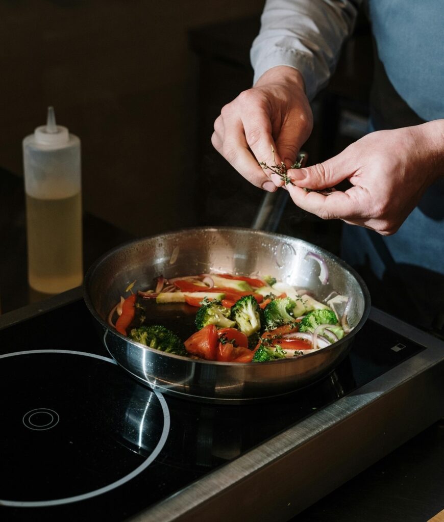 Na imagem aparece uma mão0 segurando uma erva, para colocar nos legumes que estão na frigideira, onde aparecem, brócolis, pimentão vermelho, cebola, tomate e cenoura.