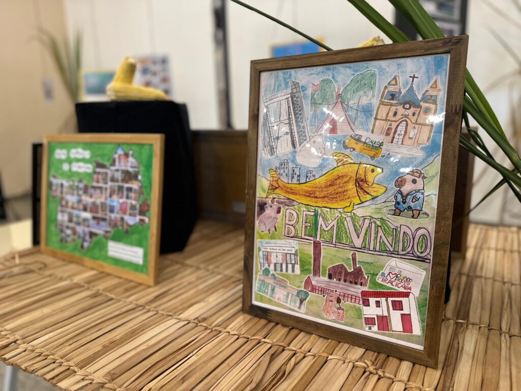 A imagem exibe uma exposição de arte em um ambiente interno, possivelmente uma galeria ou escola. Vários painéis pretos verticais estão dispostos no espaço, exibindo uma variedade de trabalhos artísticos, incluindo desenhos, fotografias e colagens. Em primeiro plano, há uma mesa com uma decoração temática, apresentando espigas de milho sobre blocos pretos e um quadro com uma paisagem de cachoeira e elementos da natureza. A decoração inclui esteiras de material natural, como bambu ou palha, que complementam a apresentação. Ao redor dos painéis, há plantas altas e secas, semelhantes a cana-de-açúcar, adicionando um toque orgânico à exposição. A iluminação é feita por spots no teto, destacando as obras de arte. O chão é de cimento polido, e as paredes são de cor clara, criando um fundo neutro para as exposições. A disposição dos painéis e a presença dos elementos decorativos sugerem um evento cultural ou uma mostra de trabalhos escolares, possivelmente com foco em temas rurais ou naturais, dada a presença do milho e das plantas.