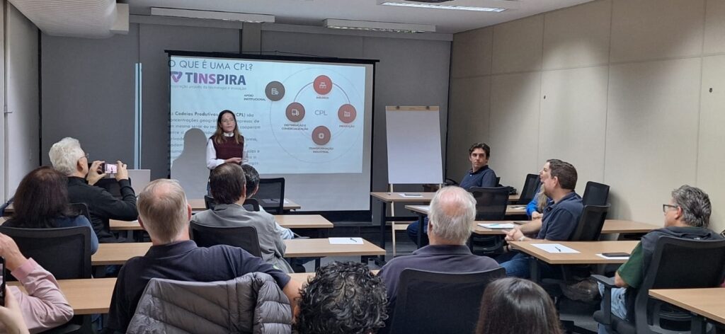 A imagem mostra uma sala de aula ou sala de treinamento onde uma mulher está apresentando um slide. O slide, que está projetado em uma tela, tem o título "O QUE É UMA CPL?" e o logotipo "TINSPIRA" com o subtítulo "Inspiração através da tecnologia e inovação". O slide também exibe um diagrama circular com o título "CPL" no centro, rodeado por quatro ícones e legendas: "INSUMOS", "PRODUÇÃO", "TRANSFORMAÇÃO INDUSTRIAL" e "DISTRIBUIÇÃO E COMERCIALIZAÇÃO". Há também um ícone e legenda para "APOIO INSTITUCIONAL" fora do círculo. A apresentadora, vestida com uma blusa branca e um colete vinho, está em pé ao lado da tela, gesticulando enquanto fala. O público está sentado em mesas em frente à tela, com alguns indivíduos tirando fotos da apresentação com seus celulares. A sala parece ser moderna, com iluminação embutida no teto e paredes revestidas com painéis. O ambiente sugere uma sessão de aprendizado ou compartilhamento de conhecimento.