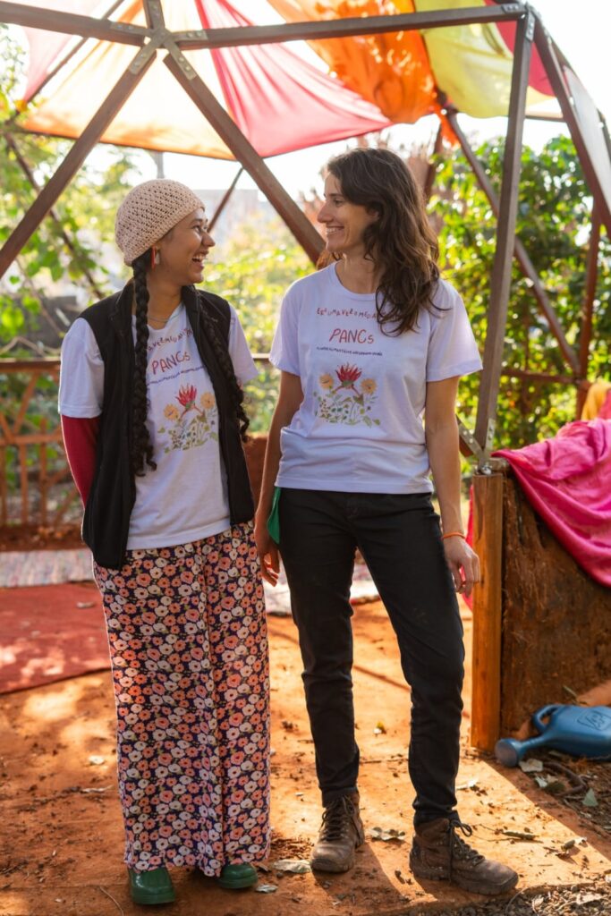 A imagem mostra duas mulheres em pé, sorrindo uma para a outra, dentro de uma estrutura semelhante a uma tenda geodésica. O teto da estrutura é feito de tecidos coloridos em tons de laranja, amarelo e vermelho, que filtram a luz do sol, criando um ambiente acolhedor. A mulher à esquerda usa um gorro de crochê bege, um colete preto sobre uma camiseta branca com uma estampa de flores e plantas, e uma calça comprida com um padrão floral em tons de rosa e laranja sobre um fundo azul escuro. Ela tem tranças longas e usa um par de sapatos verdes. A mulher à direita tem cabelos castanhos longos e ondulados e veste uma camiseta branca com a mesma estampa de flores e plantas, com a inscrição "ERA UMA VEZ O MEU AMOR PANCS". Ela usa calças pretas justas e botas marrons desgastadas. O chão é de terra batida e há vegetação ao fundo, sugerindo um ambiente ao ar livre ou um jardim. Há também um regador azul no canto inferior direito da imagem e um tecido rosa vibrante pendurado em uma das laterais da estrutura. A iluminação geral da foto é quente, realçando as cores e as texturas.