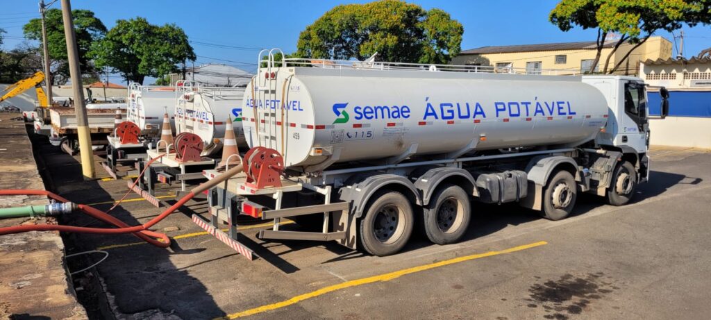 A imagem mostra uma fila de caminhões-tanque brancos estacionados em uma superfície pavimentada. Os caminhões têm "SEMAE ÁGUA POTÁVEL" escrito nas laterais, indicando que são para transporte de água potável e pertencem ao SEMAE (Serviço Municipal de Água e Esgoto). Detalhes visíveis na imagem: * *Caminhões:* Vários caminhões-tanque brancos estão alinhados. O caminhão mais próximo é um caminhão-trator com um grande tanque cilíndrico branco. * *Identificação:* O texto "SEMAE ÁGUA POTÁVEL" é exibido de forma proeminente na lateral do tanque. O logo do SEMAE, um "S" estilizado em verde, também é visível. Números como "115" e "459" estão presentes nos tanques. * *Mangueiras e Equipamentos:* Mangueiras vermelhas estão conectadas a alguns dos caminhões, com uma mangueira parecendo estar conectada a uma mangueira verde no chão, sugerindo uma operação de enchimento ou esvaziamento. Cone de sinalização laranja e carretéis de mangueira vermelha também são visíveis na parte traseira dos caminhões. * *Ambiente:* Os caminhões estão estacionados ao ar livre, com um céu azul claro e luz solar. Árvores com folhagem verde e amarela são visíveis ao fundo, junto com um edifício. O chão é pavimentado, com linhas amarelas marcando os espaços de estacionamento. * *Detalhes:* Os caminhões têm múltiplos eixos e pneus grandes. Alguns sinais de desgaste são visíveis, consistentes com veículos operacionais. Uma mancha escura, possivelmente de óleo ou combustível, é visível no pavimento em primeiro plano.