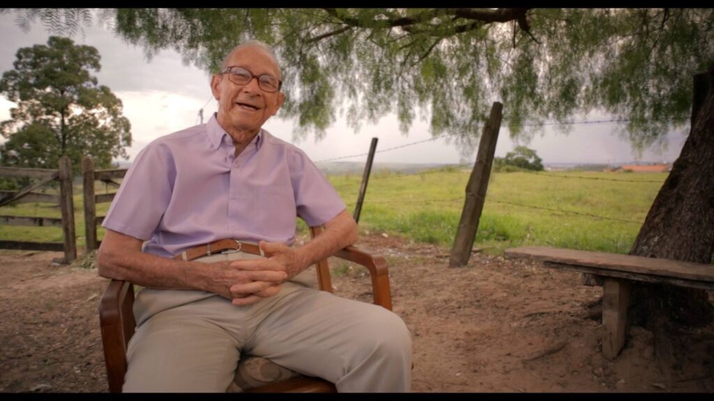 A imagem mostra um homem idoso, de óculos, sentado em uma cadeira de madeira em um ambiente externo. Ele veste uma camisa social de manga curta na cor lilás e calças bege. As mãos dele estão cruzadas no colo, e ele sorri para a câmera. O fundo da imagem sugere um ambiente rural. Há uma cerca de madeira à esquerda, e uma paisagem com grama verde e algumas árvores ao fundo. Uma árvore com folhagem densa cobre parte do topo da imagem, criando uma sombra sobre o homem. Um banco de madeira rústico está posicionado ao lado de uma árvore maior à direita. O céu está nublado, dando um tom suave à cena.